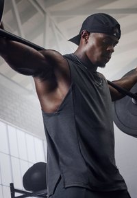T-shirt de sport sans manches noir avec une coupe ample, un tissu texturé et des fentes latérales. Individu levant une barre dans une salle de sport.