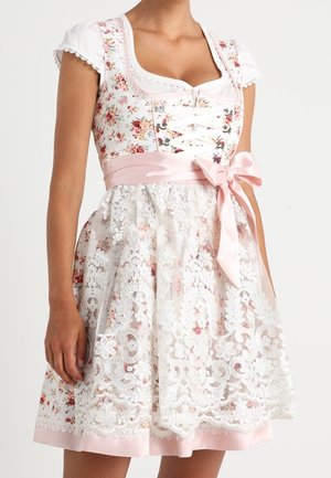Kvinde iført en hvid blomsterdirndl kjole med blondeforklæde og lyserød satin sløjfe bundet i taljen, korte puffede ærmer.