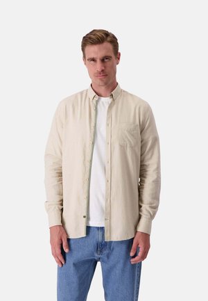 Camicia beige a maniche lunghe con colletto botton-down, taschino sul petto, leggera texture, indossata sopra una t-shirt bianca, abbinata a jeans blu.