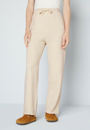 Beige, soft fabric joggers with a drawstring waistband and frayed hem. Loose fit. Pair of brown suede slippers visible at the bottom.