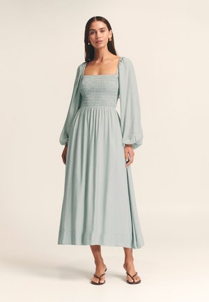 Hellblaues, langärmliges Maxikleid mit quadratischem Ausschnitt und gerafftem Oberteil. Fließender Stoff mit subtiler Textur und gespaltenem Saum.