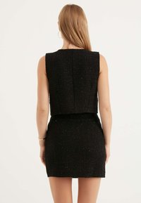 Svart tweedklänning med en figursydd silhuett, ärmlös design och kort topp. Har ett texturerat tyg med subtila glittrande detaljer.