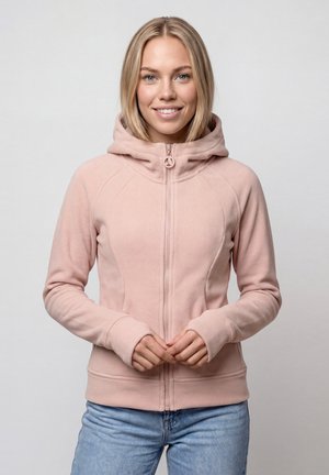 Junge Frau lächelt, trägt einen hellrosa Zip-up-Hoodie und blaue Jeans, steht vor einem schlichten hellen Hintergrund.