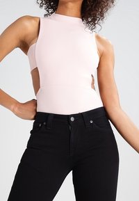 Bodysuit recortado en rosa suave con cuello alto y diseño sin mangas, combinado con jeans negros de tiro alto con bolsillos delanteros.