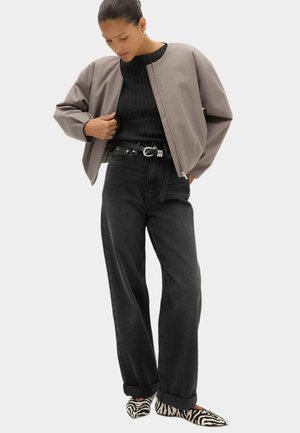 Kvinde iført taupe cropet jakke, sort ribstrikket sweater, højtaljede sorte jeans med bælte og zebra-print flade sko, ser ned og justerer jakken.