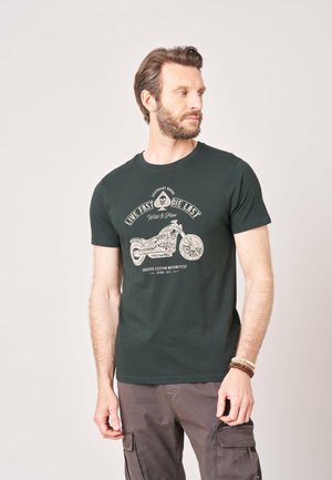 Homme avec barbe portant un t-shirt noir à motif moto et un pantalon cargo, regardant sur le côté devant un fond clair uni.