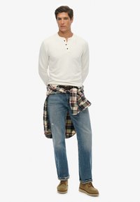 Chemise à manches longues blanche en henley avec trois boutons, jeans bleu clair, et flanelle à carreaux rouge et noir nouée à la taille. Chaussures beiges.