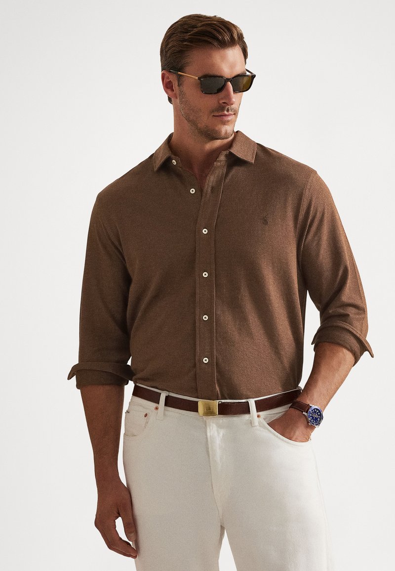 Ά�νδρας που φοράει καφέ υφασμάτινο πουκάμισο με κουμπιά και μανίκια γυρισμένα, λευκό παντελόνι, καφέ ζώνη, μπλε ρολόι και γυαλιά ηλίου, στέκεται με το ένα χέρι στην τσέπη.