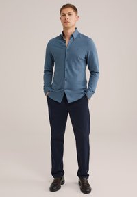 Garā piedurkņu pogājamā krekls teāl krāsā, izgatavots no gludas auduma, apvienots ar tumši ziliem biksēm un melniem loafers, rokas kabatās.