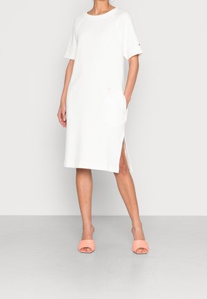 Kobieta ubrana w luźną, białą sukienkę do kolan z bocznym rozcięciem i krótkimi rękawami, w połączeniu z różowymi, otwartymi klapkami na palce.