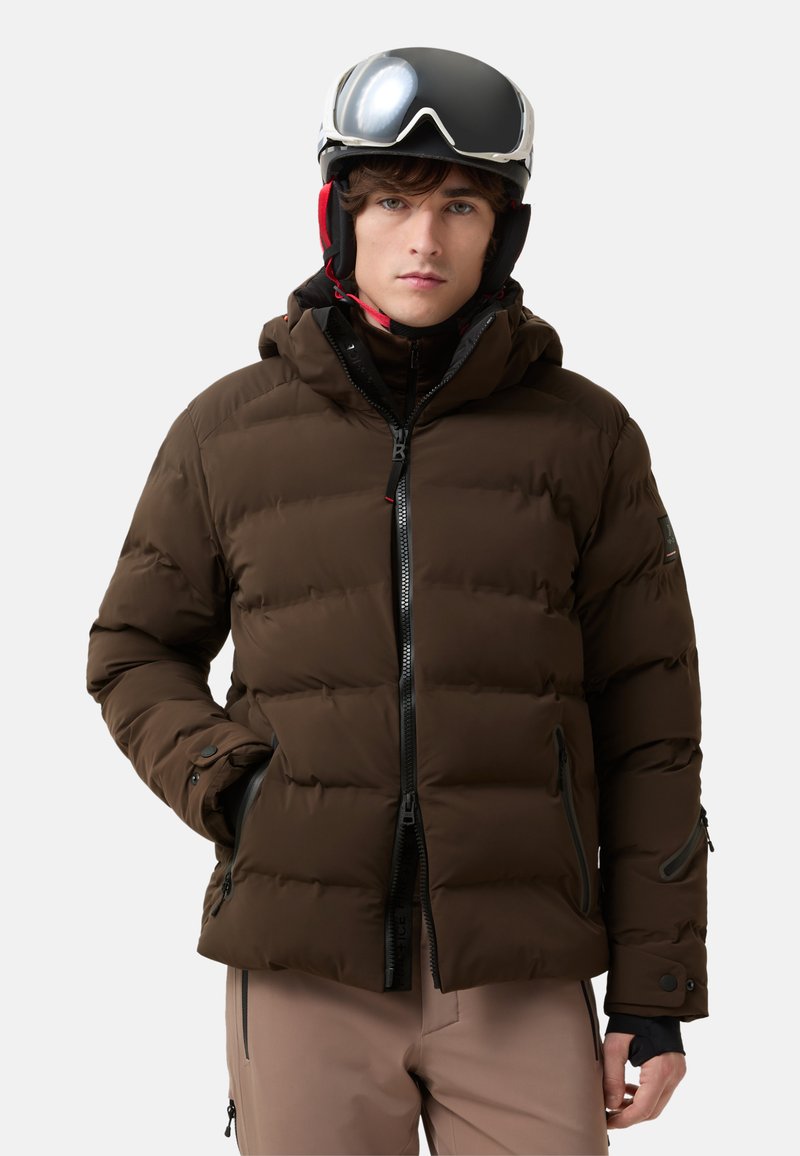 Young person wearing a brown padded winter jacket, beige pants, and a black ski helmet with goggles against a plain background.