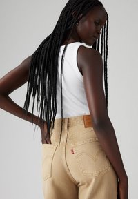 Μπεζ τζιν ψηλόμεσα με δερμάτινο ταμπελάκι σε ταμπά χρώμα στο πίσω μέρος, συνδυασμένα με εφαρμοστό λευκό ριμπ κορμάκι. Μακριά μαύρα πλεξούδες.