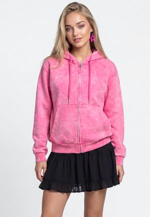 Ung kvinde iført en pink lynlåshoodie, sort flæset nederdel og store lyserøde stjerneformede øreringe, stående på en ensfarvet hvid baggrund.