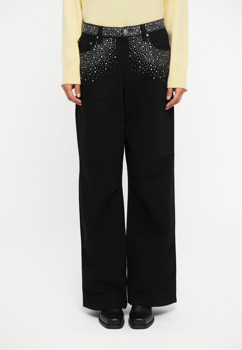 Wide-leg black trousers featuring a studded accent on the waistband, crafted from denim with a relaxed fit. Styled with black flat shoes.