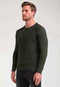 Maglione lavorato a maglia verde scuro con un motivo geometrico testurizzato, scollatura rotonda e polsini aderenti. Indossato con jeans scuri.