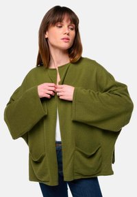 Femme aux cheveux bruns portant un cardigan olive décontracté à manches larges et poches avant, le fermant sur un haut blanc et un jean bleu.