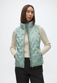 Mentolovo zelená, prešívaná vesta s vysokým golierom a predným zipsom. Obsahuje bočné vrecká a nosí sa cez krémový rebrovaný sveter.