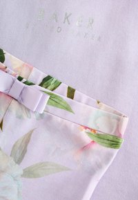 Tissu floral avec des feuilles vertes et des fleurs roses pâles, nœud lavande attaché, sur un tissu violet clair avec le texte "Baker by Ted Baker".