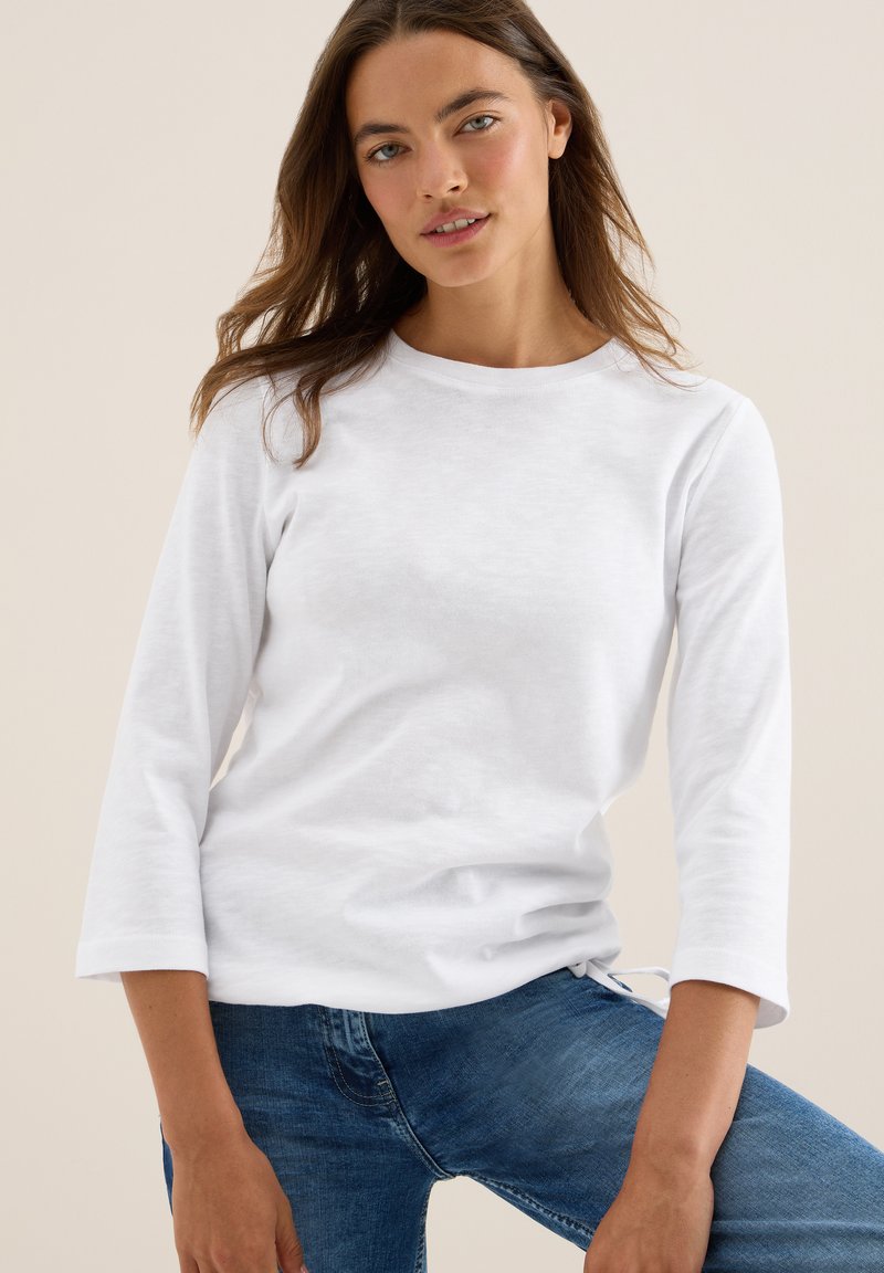 Chemise blanche à manches longues avec un col rond et une texture douce. Associée à un pantalon en denim bleu, montrant un style décontracté et ajusté.