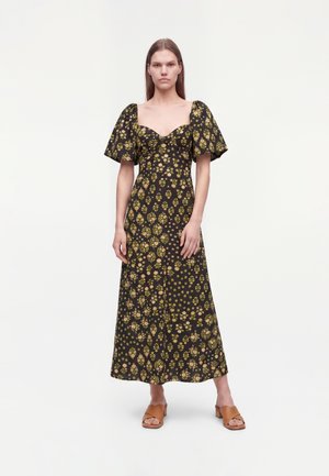 Vrouw draagt een zwarte enkellange jurk met groene en gele bloemenpatronen en opgeblazen korte mouwen, gecombineerd met tan open-toe sandalen.