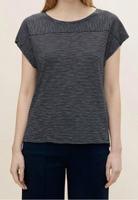 Kvinna iklädd en svartvit randig kortärmad topp och mörka byxor, ståendes mot en enhetlig ljus bakgrund.