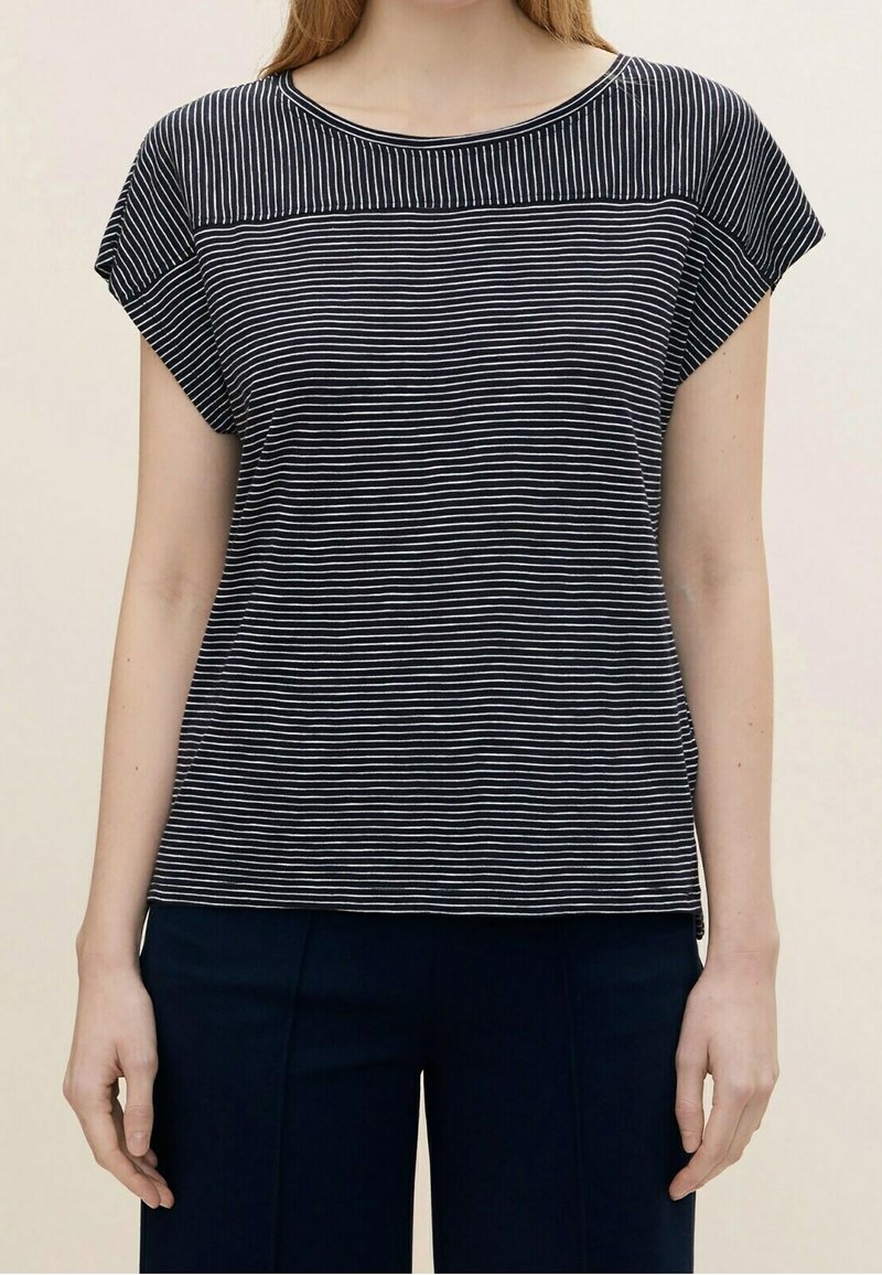 Kvinna iklädd en svartvit randig kortärmad topp och mörka byxor, ståendes mot en enhetlig ljus bakgrund.