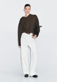 Brauner übergroßer Strickpullover mit V-Ausschnitt und seitlicher Knotendetail, kombiniert mit weißen weit geschnittenen Hosen und schwarzen Holzclogs. Sanfte Texturen, lässige Passform.