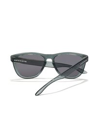 Occhiali da sole in acetato grigio con lenti scure, montatura leggermente arrotondata e dettagli neri sulle astine con il logo "HAWKERS" su un lato.