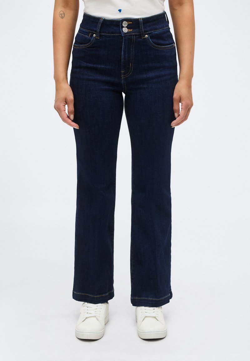 Σκούρα μπλε τζιν με ίσια γραμμή, ψηλή μέση, διπλό κουμπί και κλείσιμο με φερμουάρ, συνδυασμένα με λευκά αθλητικά παπούτσια και λευκό τοπ.