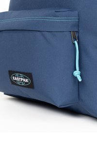 Blauer Stoffrucksack mit einer Vorderetasche, die einen türkisfarbenen Reißverschlussanhänger, ein schwarzes Logopatch und kontrastierende Nähte aufweist. Strukturierte Oberflächendetails.