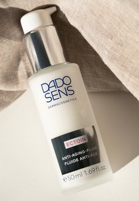 Weiße Flasche mit silberner Pumpdeckel, beschriftet mit "DADO SENS DERMACOSMETICS ECTOIN ANTI-AGING-FLUID", auf einem weichen beigefarbenen Stoff.