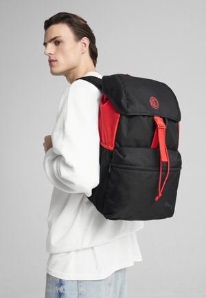 Zaino nero e rosso con chiusura a patta, tracolla regolabile, tasca con zip e tasche laterali in rete. Presenta un logo sul davanti.