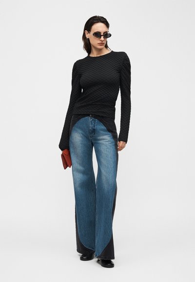 Γυναίκα που φοράει μαύρο μακρυμάνικο τοπ με υφή, τζιν παντελόνι με φαρδύ πόδι σε δύο αποχρώσεις, μαύρες μπότες, γυαλιά ηλίου και κρατάει ένα μικρό κόκκινο τσαντάκι.