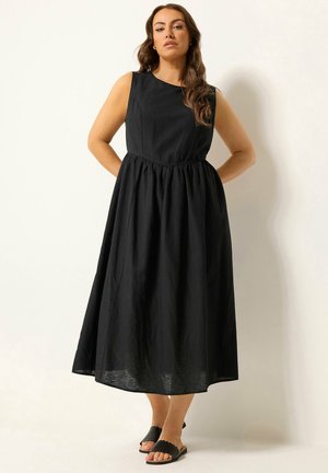 Kobieta stoi, ubrana w czarną sukienkę midi bez rękawów z marszczoną talią i czarne sandały wsuwane na tle jednolitego jasnego tła.