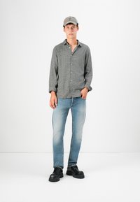 Camisa de manga larga a cuadros en gris, jeans de mezclilla azul claro y zapatos de suela gruesa negros; combinados con una gorra gris, de pie contra un fondo blanco.