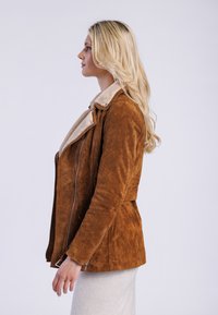 Veste en daim marron avec un col en shearling crème, dotée d'une fermeture éclair inclinée et d'une coupe ajustée, présentée de profil sur un fond neutre.