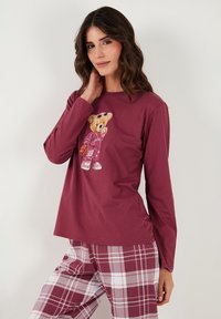 Bordó hosszú ujjú felső maci grafikával, piros-fehér kockás nadrággal párosítva. Puha anyag, kényelmes szabás, kerek nyakkivágás.