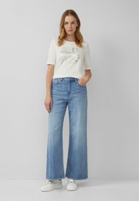 T-shirt blanc avec un design abstrait métallique, associé à un jean large bleu clair. Baskets blanches. Style simple et décontracté.
