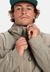 Beige waterdichte jas met een hoge kraag, ritsen en verstelbare manchetten. Beschikt over een gestructureerde stof en een trekkoord bij de halslijn.