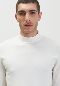 Maglione bianco a maglia con colletto alto, texture liscia e design aderente. Lo stile minimalista enfatizza la semplicità e la modernità.