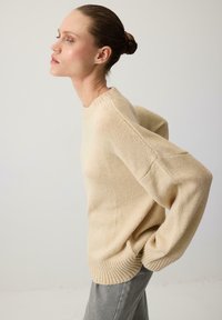 Béžový sveter s okrúhlym výstrihom, spadnutými ramennými švami a rebrovaným lemom. Mäkka štruktúra a voľný strih; skombinovaný so šedými nohavicami.