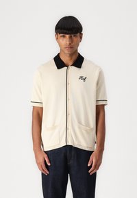 Kurzarmpullover in Creme mit einem schwarzen Kragen und Bündchen. Mit Knopfleiste und zwei vorderen Taschen. "Huf" auf der Brust gestickt.