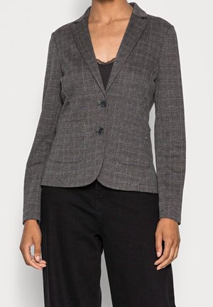 Kvinde iført en tætsiddende grå ternet blazer med to sorte knapper over en sort blonder-trimmet top, kombineret med sorte bukser.