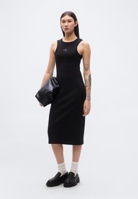 Μαύρο μίντι φόρεμα χωρίς μανίκια με λογότυπο, εφαρμοστή σχεδίαση, λεία υφή. Συνδυασμένο με μαύρο clutch και στιλιστικά λευκές κάλτσες σε στρώσεις και μαύρα παπούτσια slip-on.