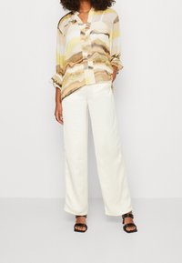 Vrouw in losse witte broek, open-kraag beige, bruin en gele blouse met patroon en zwarte haksandalen, staande tegen een effen achtergrond.