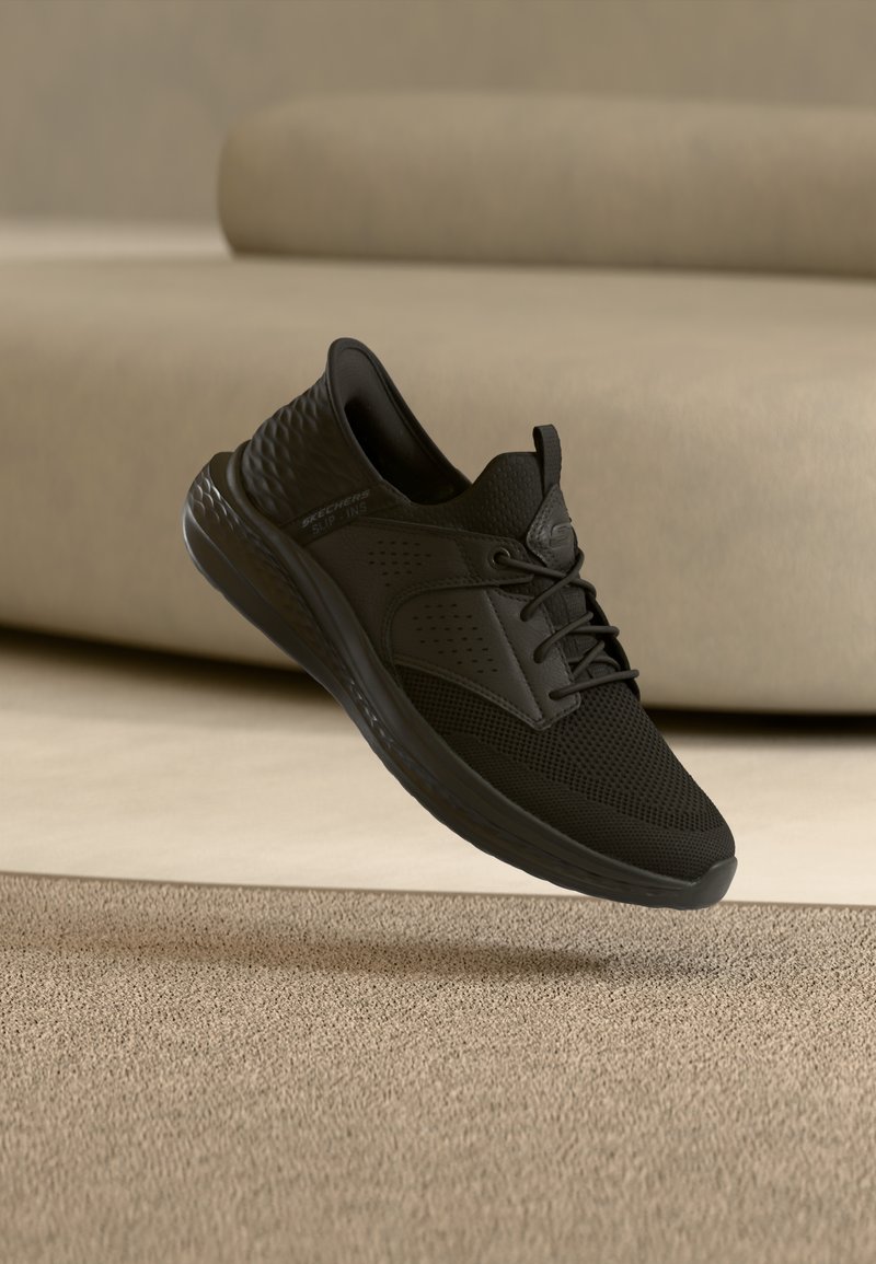 Μαύρο αθλητικό παπούτσι με επάνω μέρος από δίχτυ και κορδόνια να αιωρείται πάνω από μπεζ χαλί μπροστά από μπεζ καναπέ.