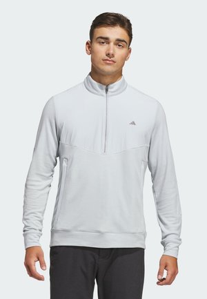 Junger Mann trägt einen hellgrauen langärmligen Quarter-Zip-Pullover mit Seitentaschen und dunkle Hose, steht mit Blick nach vorne.