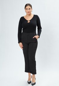 Schwarzes langärmliges Oberteil mit einem Schlüssellochdetail am Ausschnitt, kombiniert mit maßgeschneiderten schwarzen Hosen und schwarzen Schuhen mit Absatz und dekorativen Akzenten.