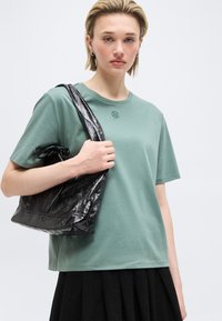 Νέα γυναίκα με κοντά ξανθά μαλλιά φοράει πράσινο μπλουζάκι και κρατά μια λαμπερή μαύρη τσάντα ώμου σε ένα απλό φόντο.