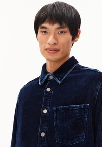 Navy corduroy shirt met een kraag, zilveren knopen en een borstzak. Opvallende ribbeltextuur en contrasterende halsafwerking.